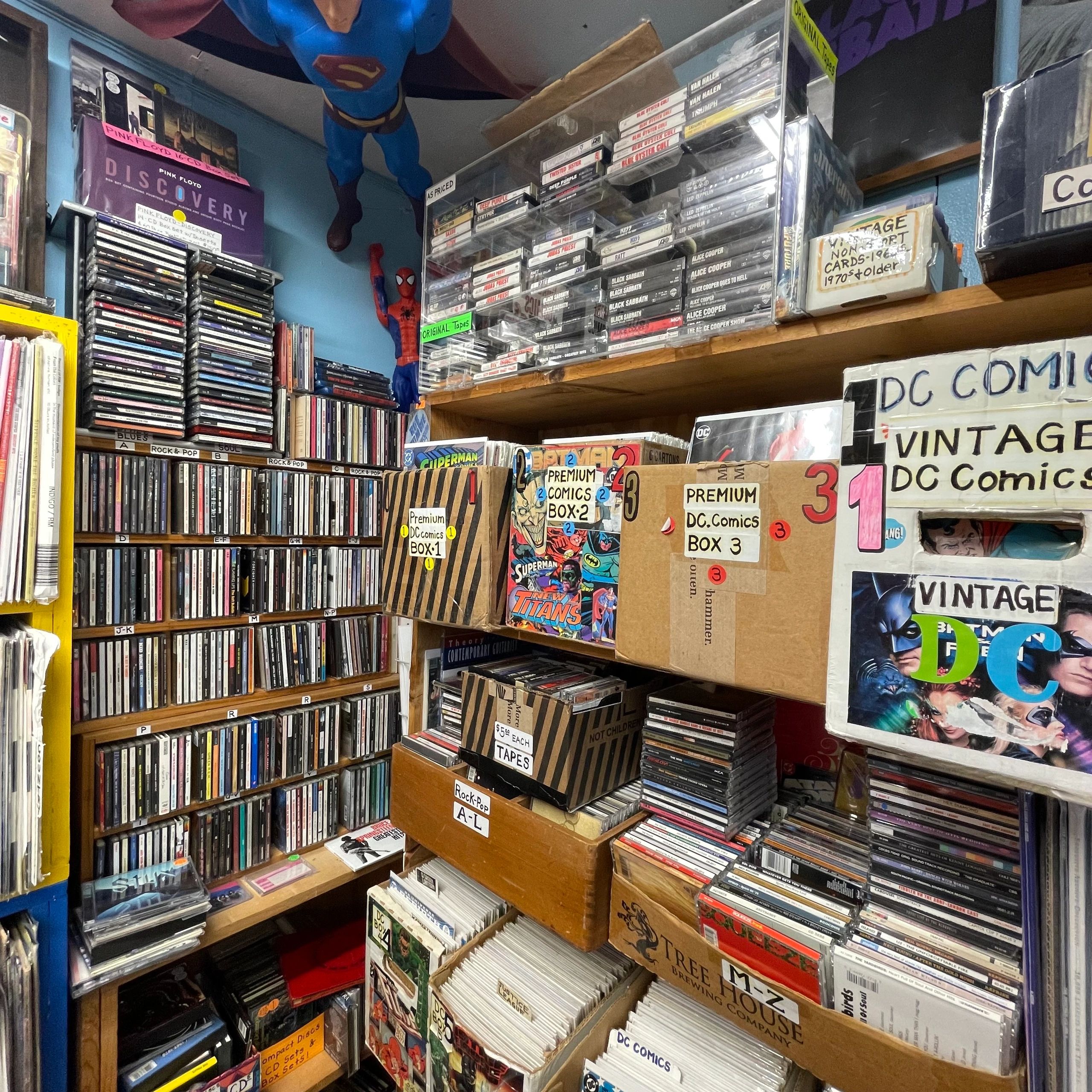 A cluttered comic and media store corner with vintage DC comics and assorted tapes and CDs